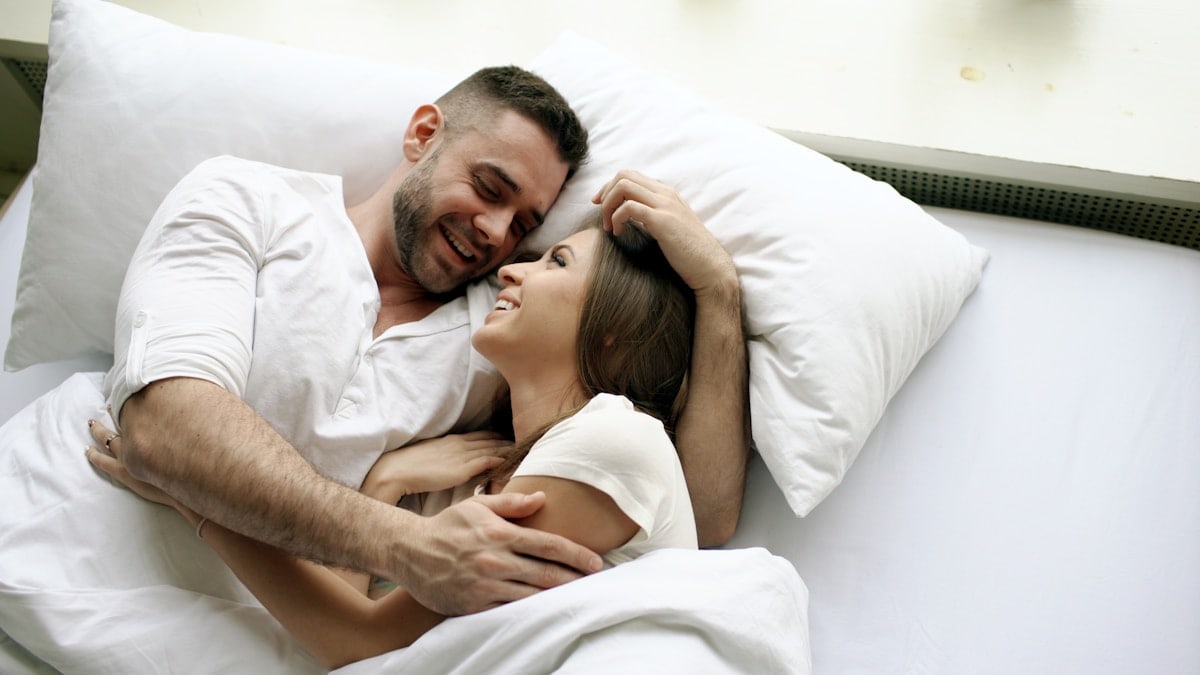 Young beautiful and loving couple kiss and hug into bed while waking up in the morning